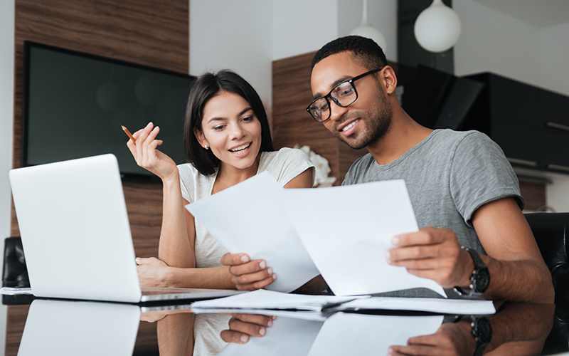 Photo of cheerful loving young couple using laptop and analyzing their finances with documents.